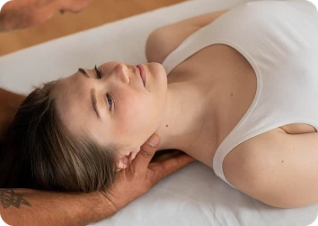 Young woman in a white tank top lying on a treatment table as a therapist supports her neck with both hands.