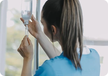 Female healthcare professional in blue scrubs adjusting an IV drip bag near a bright window.