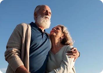 Elderly couple embracing outdoors under a clear blue sky, the woman gazing up affectionately at her bearded partner.