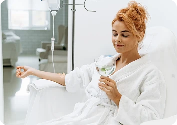 Red-haired woman in a white bathrobe relaxing during IV therapy while sipping water in a bright clinic.