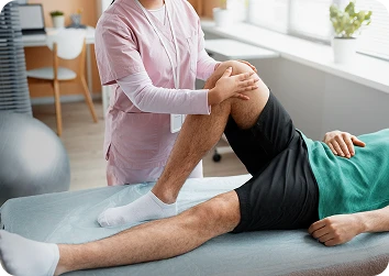 Female physiotherapist in pink scrubs guides a male patient’s bent knee during a rehabilitation exercise on a treatment table in a bright clinic.