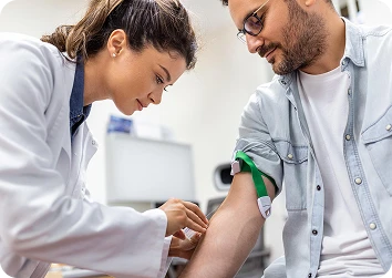 Female healthcare professional in a white coat preparing to draw blood from a seated man with a tourniquet on his arm in a bright clinic.