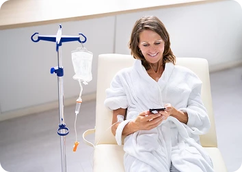 Smiling woman in a white bathrobe reclining during IV therapy while browsing her smartphone in a bright clinic.
