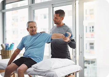 Older man in a blue T-shirt performing a seated shoulder exercise with a small dumbbell while a physical therapist guides his arm in a bright rehabilitation room.