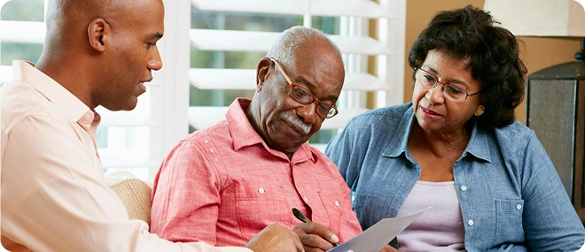 Older man in a pink shirt signs a document as an older woman listens and a male advisor explains the details in a bright living-room setting.