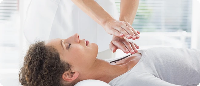 Therapist holding hands just above a woman’s chest during a Reiki-style energy healing session while the patient lies relaxed on a treatment table in a bright clinic.