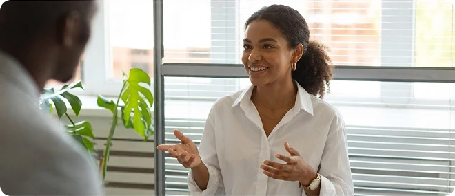 Smiling young woman in a white shirt gesturing while speaking with another person in a bright office.