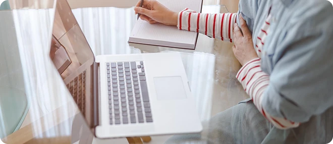 Person in a striped shirt writing in a notebook beside an open laptop on a glass table.