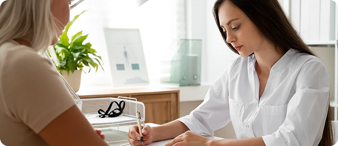 Young female therapist in a white blouse writes notes while speaking with a client across a desk in a bright office.