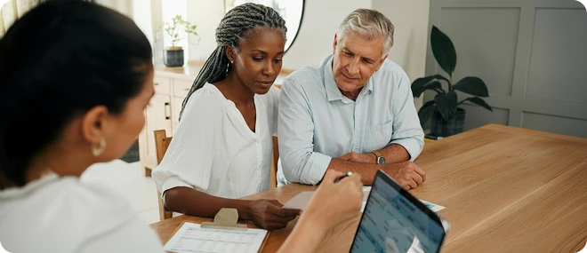 Middle-aged interracial couple reviewing paperwork with a consultant who shows information on a laptop at a wooden table in a bright home office.