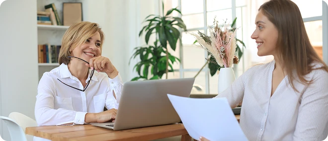 Smiling middle-aged woman holding her glasses sits at a laptop, chatting with a younger woman who is holding papers across a wooden desk in a bright, plant-filled office.