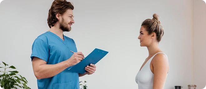 Male healthcare professional in blue scrubs holding a clipboard speaks with a female patient in a white sports top in a bright clinic.