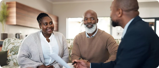 Smiling couple sitting on a sofa talking with a male advisor who holds a document in a comfortable living-room setting.