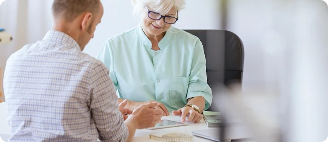 Older female healthcare professional in a mint-green coat reviews information on a tablet with a male patient at a bright office desk, a measuring tape lying nearby.