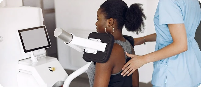 Female therapist in blue scrubs positions a young woman’s arm in a shoulder rehabilitation machine, with a monitor nearby, in a bright clinic.