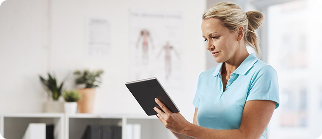 Blonde female healthcare professional in a light blue polo shirt reviewing information on a tablet in a bright clinic office.