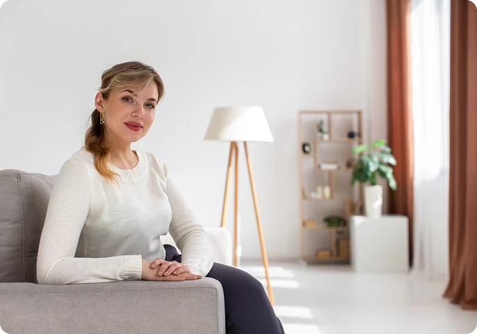 Woman with blonde hair in a light sweater sitting on a gray couch in a bright, minimalist living room, smiling calmly.