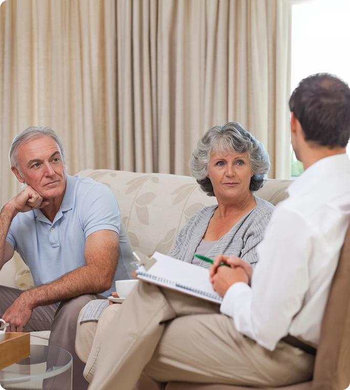 Older couple sitting on a couch listening to a male therapist with a notepad during a counseling session.
