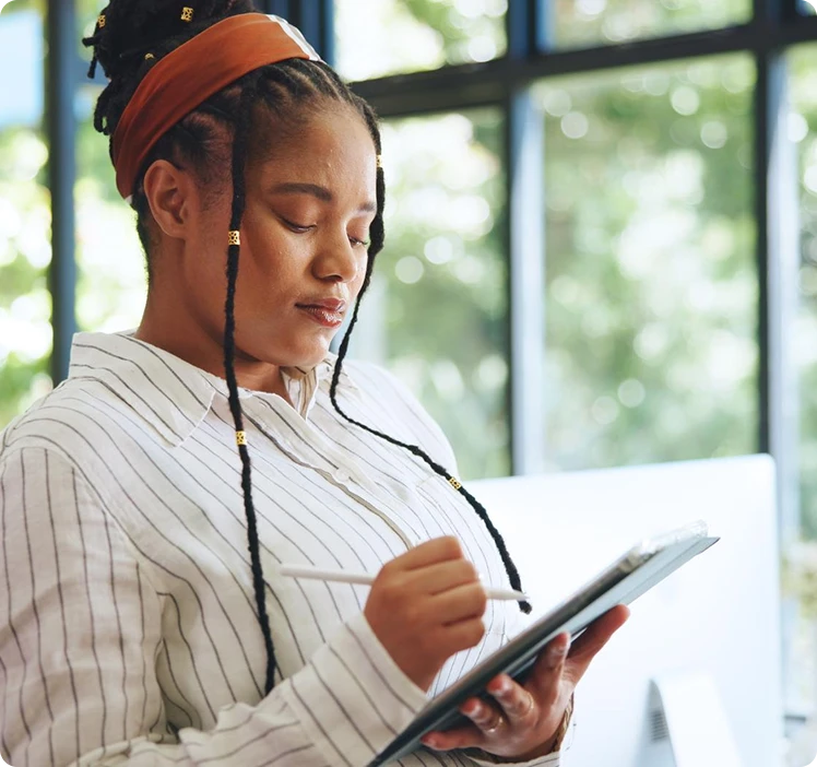 Focused young woman with braided hair and an orange headband writing on a digital tablet with a stylus in a bright office.