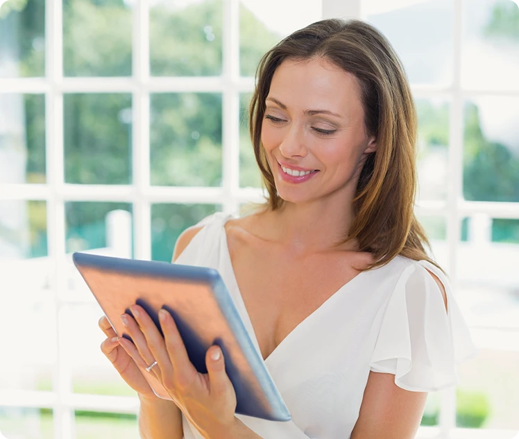 Smiling brunette woman in a white blouse holding a blue tablet near a bright window.