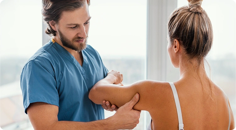 Physical therapist in blue scrubs examining a female patient's shoulder and arm during a treatment session