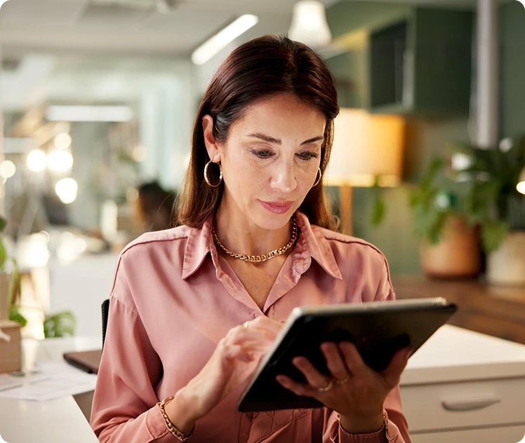 Brunette woman in a rose-pink blouse using a tablet at a modern office desk.