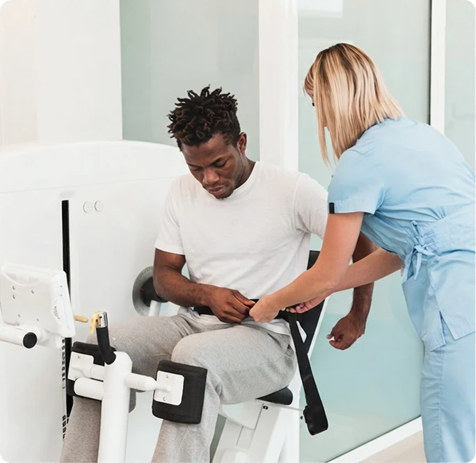 Female physical therapist in blue scrubs helping a seated male patient secure a strap on a leg exercise machine in a bright rehabilitation clinic.