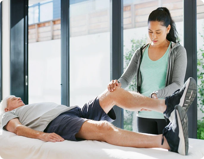 Young female physiotherapist assisting an older male patient with a leg rehabilitation exercise on a treatment table in a bright clinic.