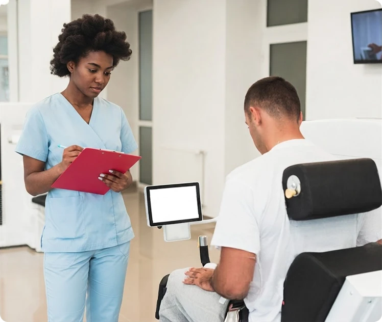 Female physiotherapist in light blue scrubs taking notes on a pink clipboard while observing a male patient using a shoulder rehabilitation machine with a blank monitor.