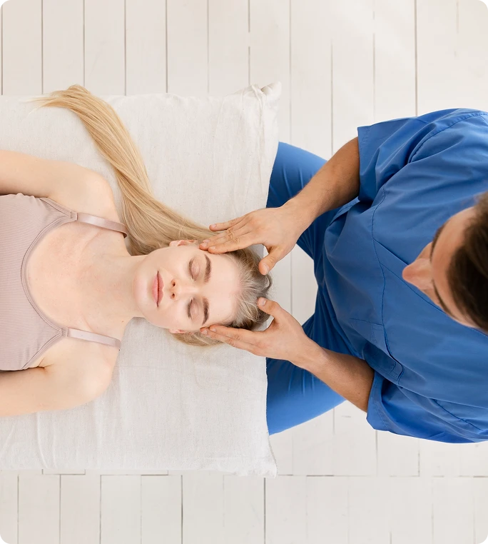 Top-down view of a therapist in blue scrubs gently massaging the temples of a relaxed woman lying on a treatment table.